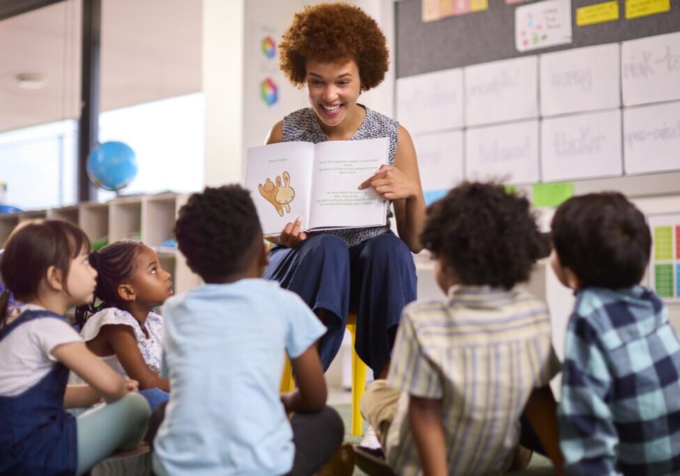 Female Teacher Reads To Multi-Cultural Elementary School Pupils Sitting On Floor In Class At School